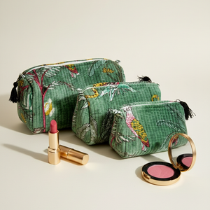 Three green patterned makeup bags with black tassels are arranged on a light beige background alongside cosmetic items including a pink lipstick, a gold compact mirror, and a pink blush.