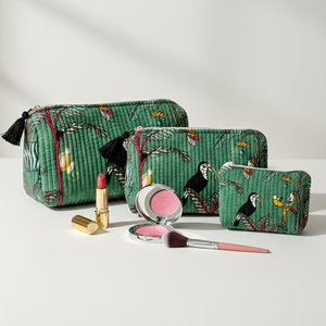 Three green quilted cosmetic pouches with a tropical toucan print are arranged on a white surface with makeup items in front, including a lipstick, compact powder, blush, and makeup brush.