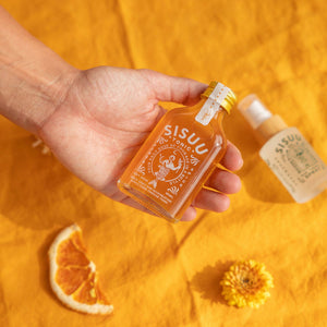 Hand holding a SISU U Tonic bottle on an orange background with oranges and flowers.