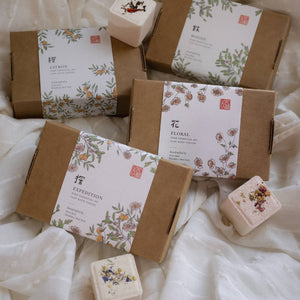 Various soap bars in brown boxes with decorative labels on a white fabric background