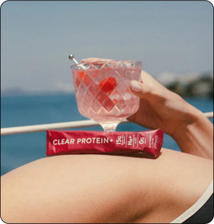 Person holding a pink cocktail with ice and a raspberry, with a protein bar in the foreground, against a blue ocean background.