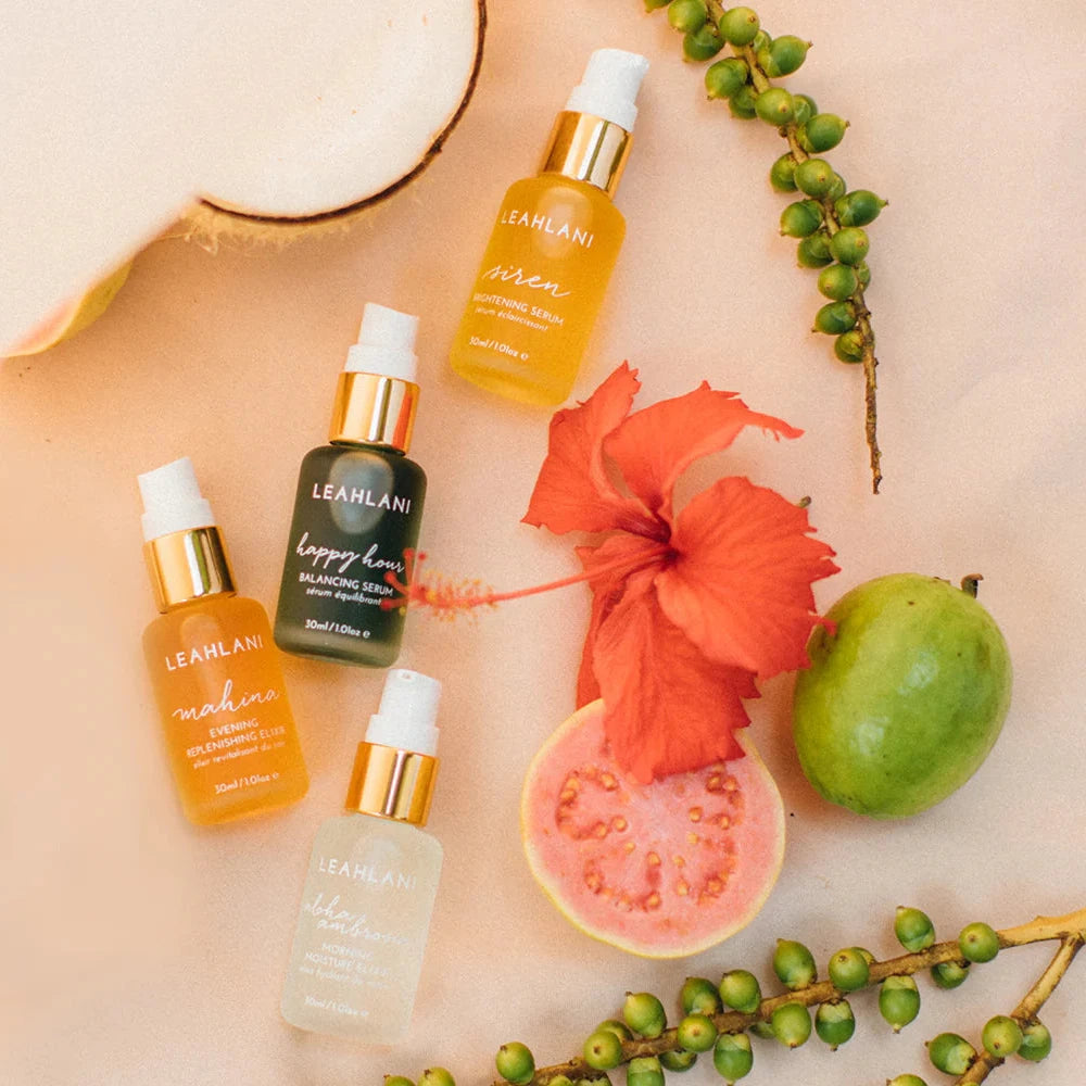 four bottles of facial oils lying next to a hibiscus flower and tropical fruit