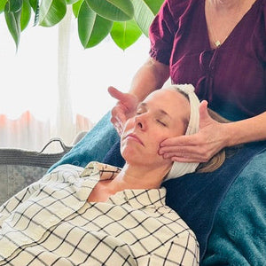 Woman giving a facial to another woman in a home setting with plants and natural light.