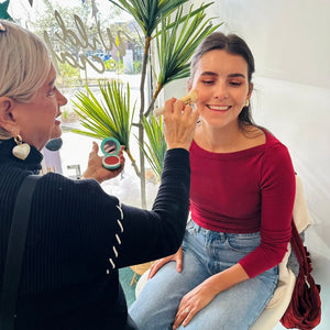 Woman getting makeup applied by another woman in a casual setting with plants and decor.