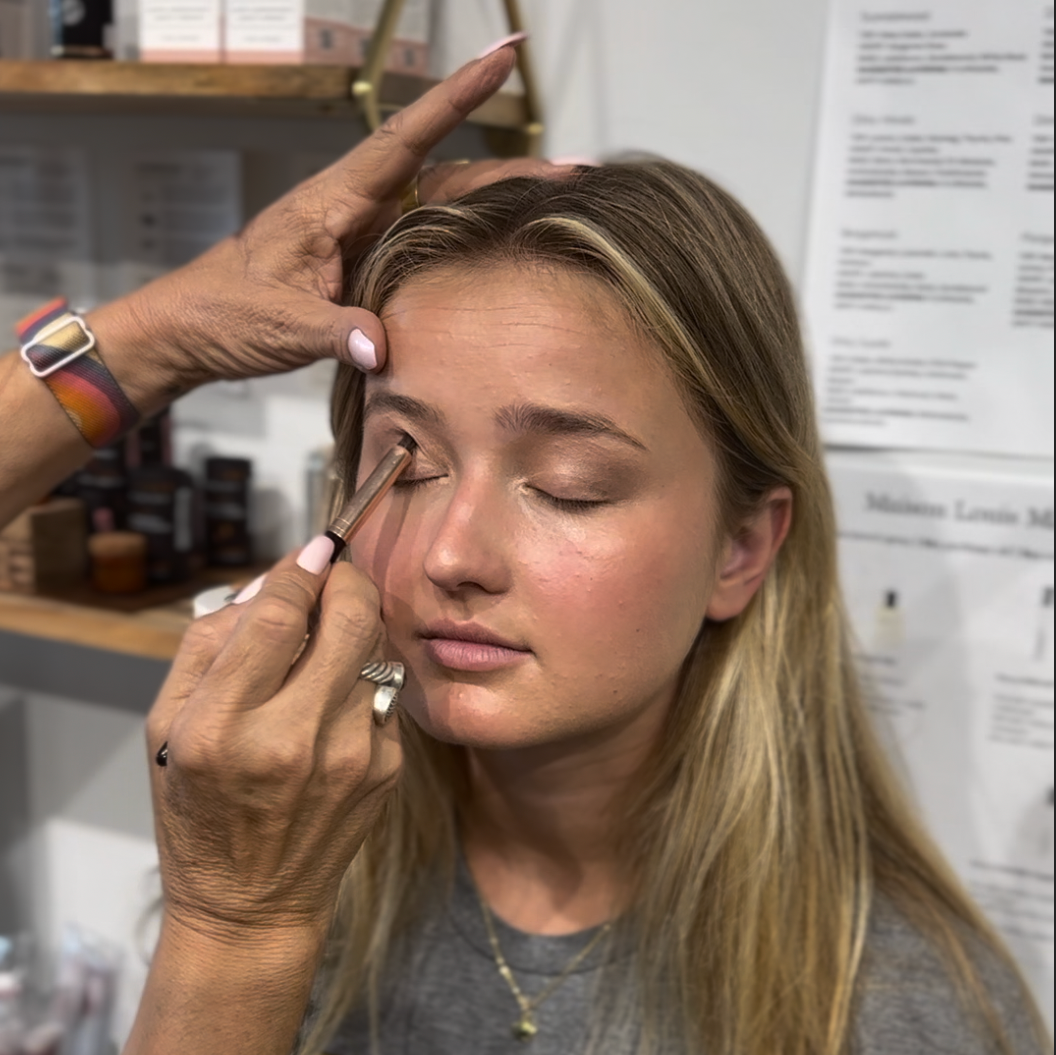 A woman receiving a makeover from an RMS Professional Makeup Artist, with the makeup artist's hands applying lipstick to the client's lips.