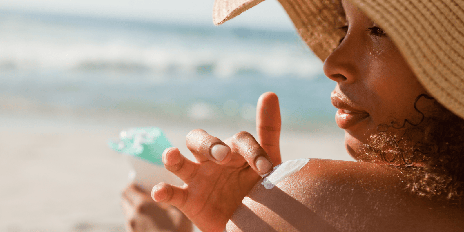 woman wearing a hat on the beach applying sunscreen to her shoulder