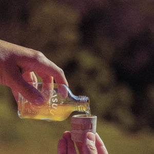 Hand pouring liquid from a small bottle into a cup with a blurred natural background