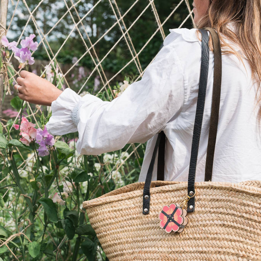 Person wearing a white jacket with a straw bag holding a pink flower charm, standing in front of a wire fence and plants.