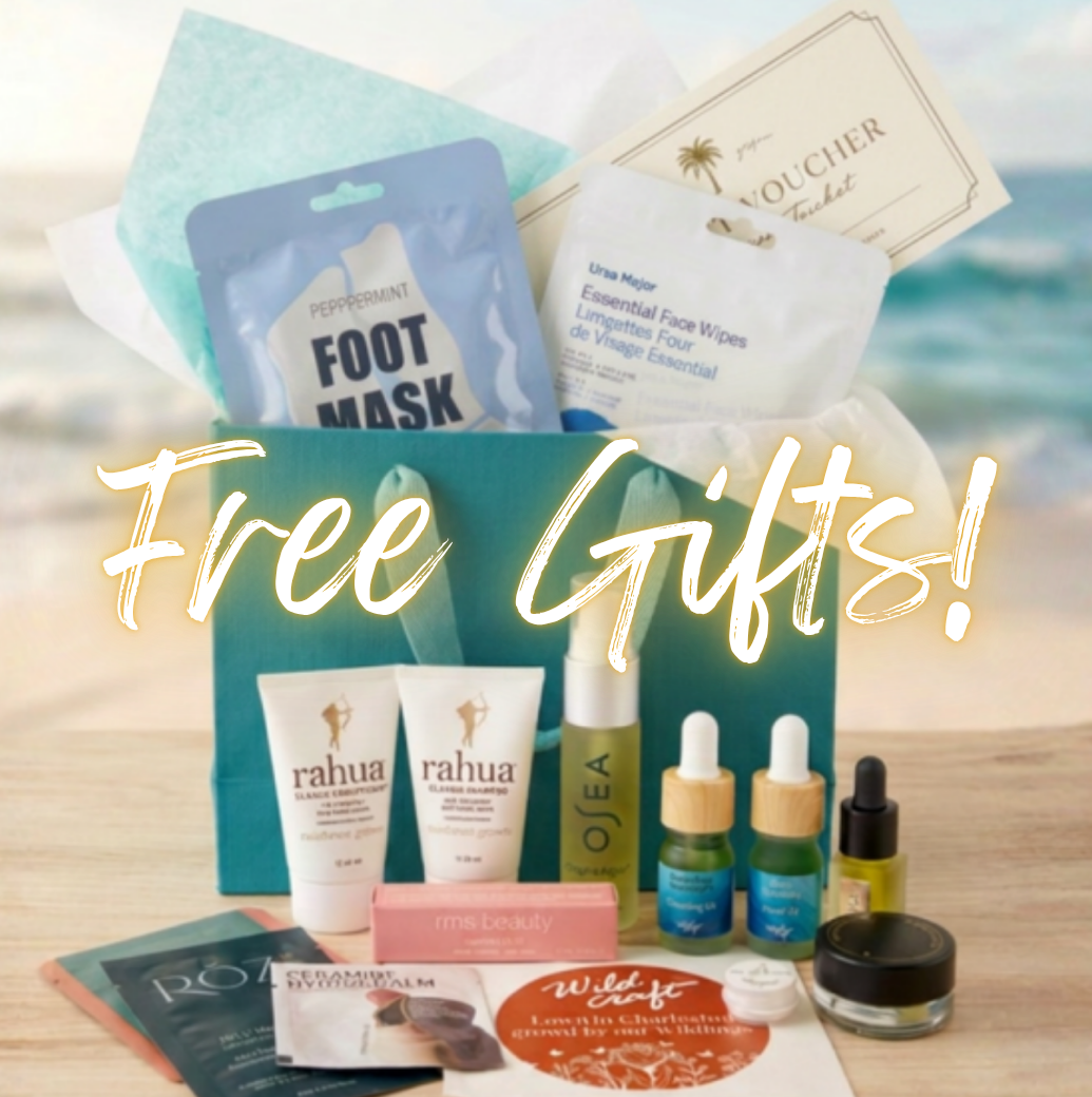 A curated beauty gift set arranged on a tabletop with a beach backdrop. Visible items include a teal gift bag, a foot mask packet, a cream face mask packet, two Rahua hand cream tubes, several small dropper bottles, a pink box, and a round jar, overlaid with the text 'Free Gifts!'.