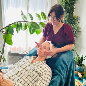 Woman giving a facial to another woman in a home setting with plants and natural light.