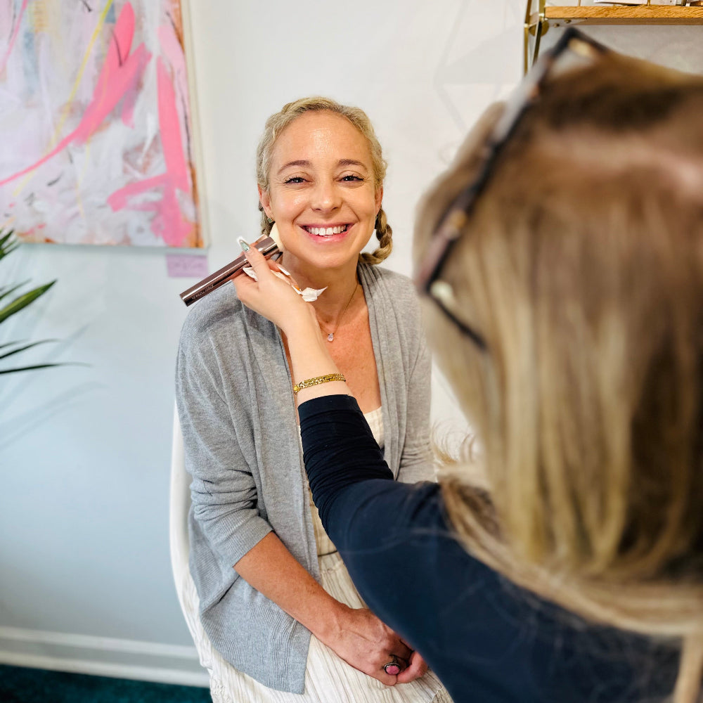 Woman getting makeup applied by another woman in a well-lit room.