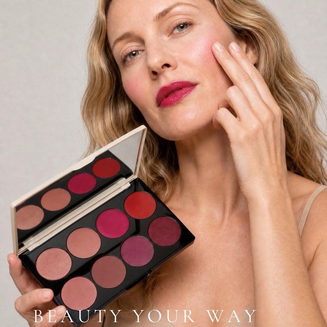 A woman with blonde hair holds an open makeup palette with multiple lip and cheek color pans, applying pink makeup to her face against a light gray background. Text at the bottom reads 'BEAUTY YOUR WAY'.