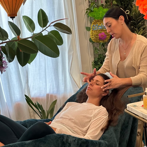 A woman receives a head and face massage while reclining on a teal massage chair in a decorated spa-like room with plants, flowers, and hanging lanterns.