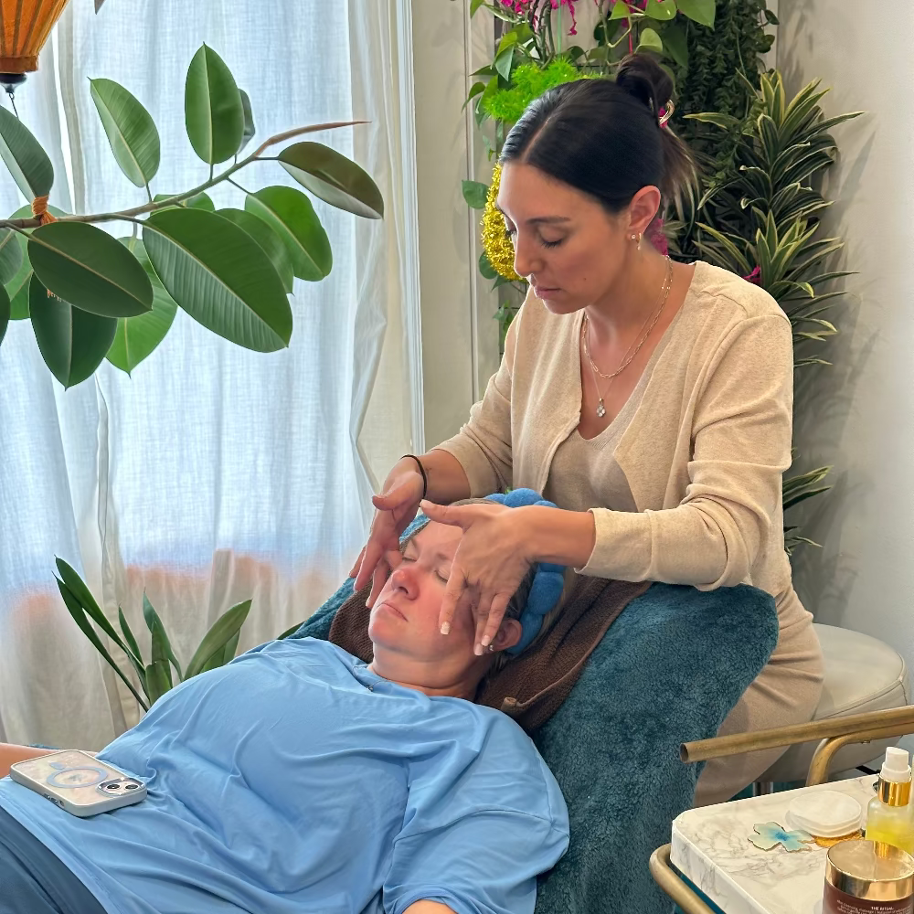 A woman receives a facial treatment while lying on a teal massage chair, with a therapist applying cream to her forehead in a bright spa-like room with plants and flowers.
